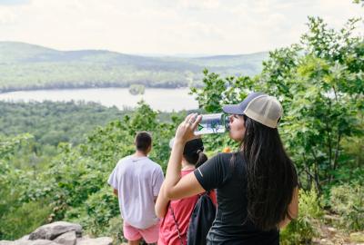 A woman with long black hair and a ball cap drinks water out of a reusable water bottle above the shores of Sebago Lake